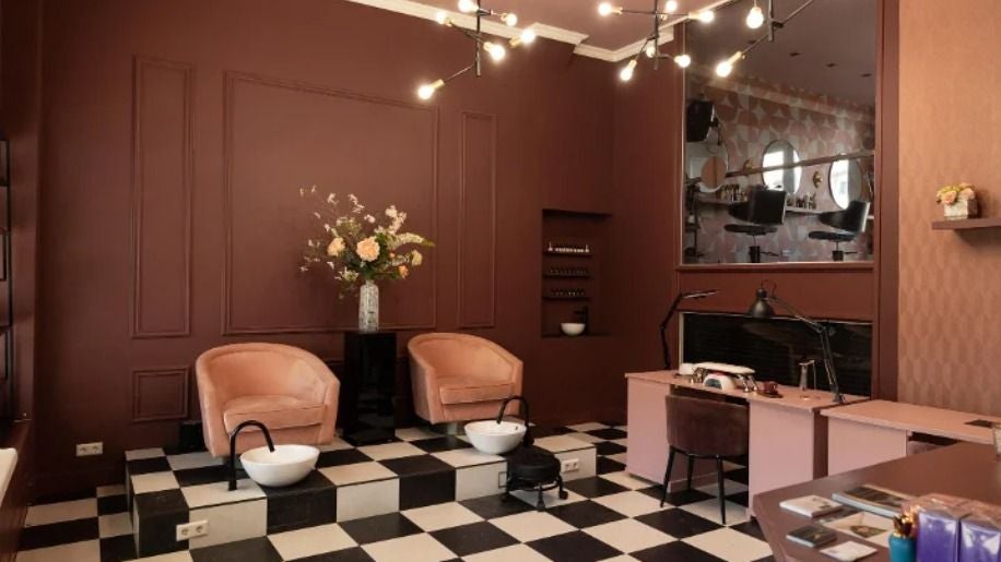 De salon: bordeauxrode wanden, een zwart-witte vloer, twee perzikkleurige pedicurestoelen en een kroonluchter
